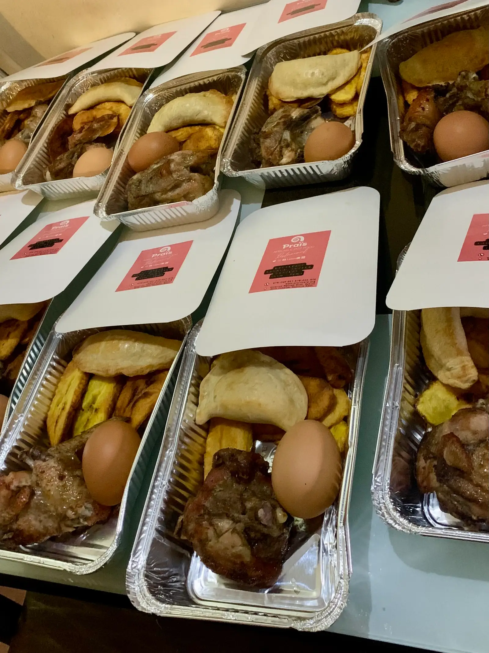 Praïs takeaway trays of plantain, fish, egg, meat and pastry, with red-labelled lids